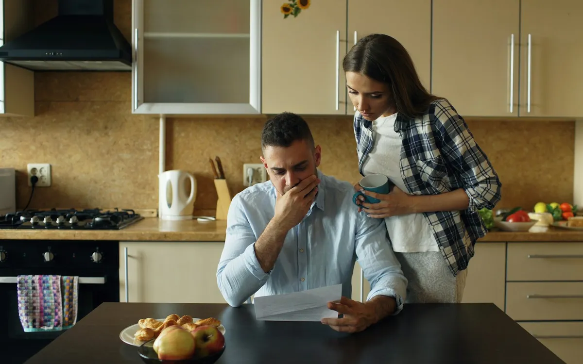 Casal preocupado na cozinha de casa, olhando um papel.